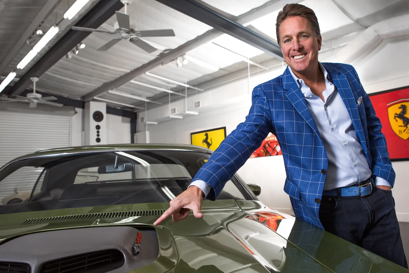 Dennis Collins in his showroom, resting a hand on the hood of a dark green muscle car, with Ferrari artwork on the wall behind him.